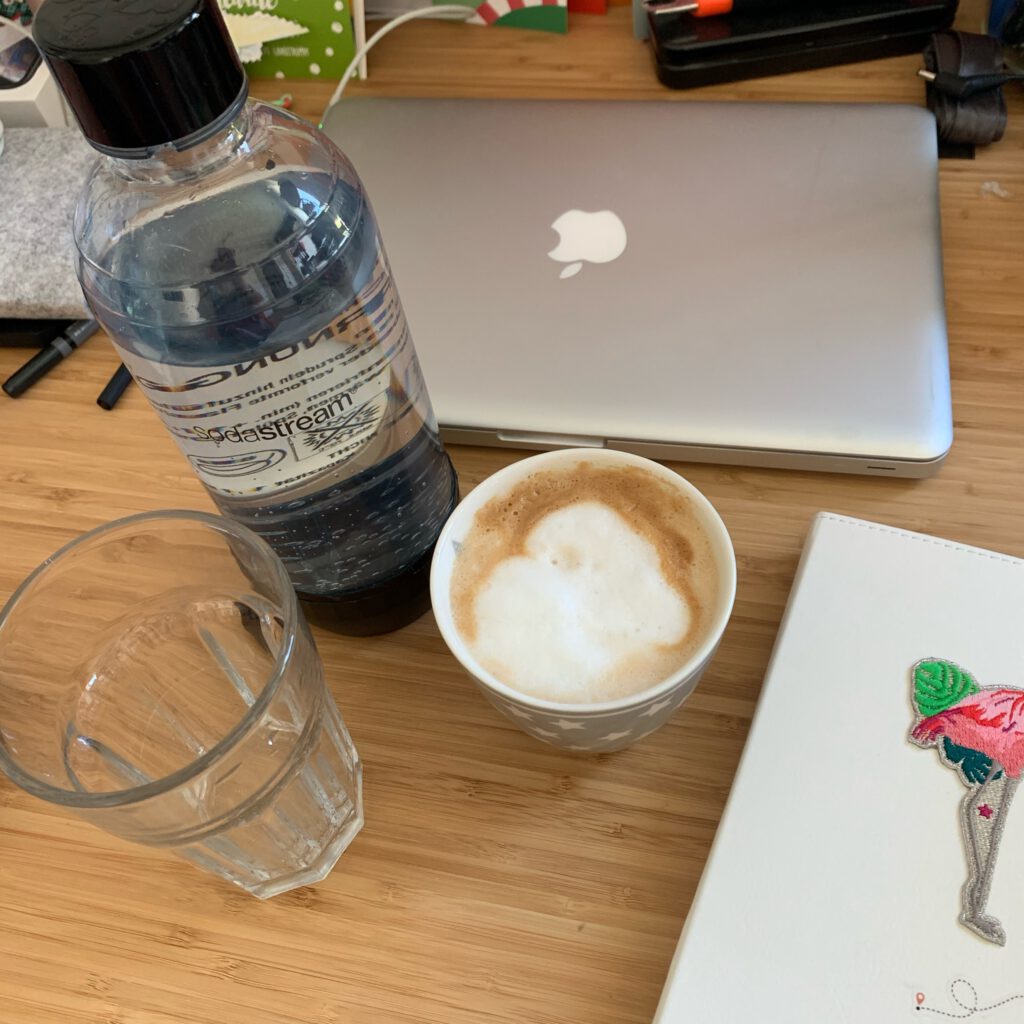 Neben einem Notebook und einem Bullet Journals stehen eine Tasse Cappuccino, eine Flasche Sprudel und ein Glase.